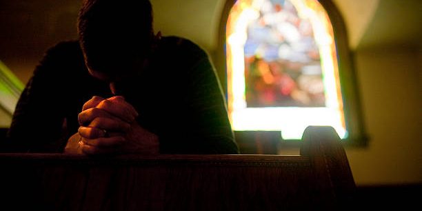 Man praying in a pew, stain glass in the background.